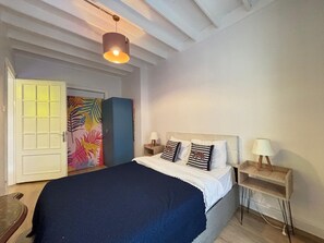 Un dormitorio con cama, mesitas de noche, lámpara colgante, un armario y una puerta decorada con una obra de arte colorida.