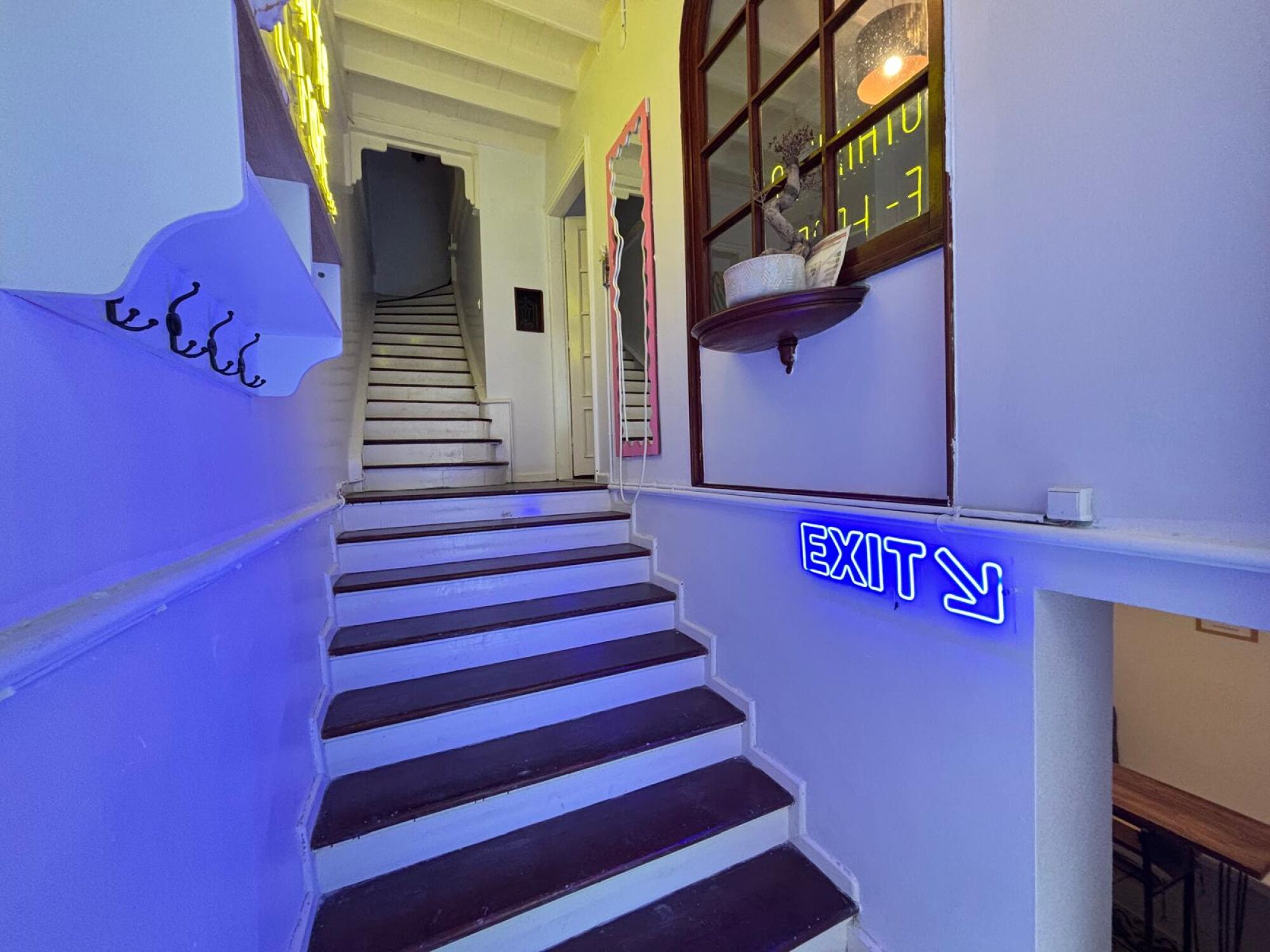 Een trap met een neon-nooduitgangbord, leidend naar een kamer met een raam en een decoratief object aan de muur.