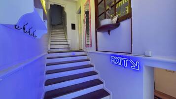 Een trap met een neon-nooduitgangbord, leidend naar een kamer met een raam en een decoratief object aan de muur.
