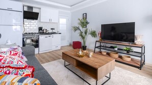 Una cocina moderna con armarios blancos y encimera negra, una mesa de centro de madera con objetos decorativos, un otoman rojo y un televisor sobre un soporte negro.