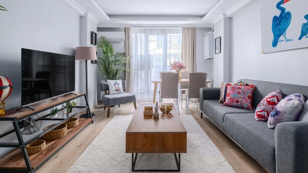 Una sala de estar moderna con un sofá gris, una mesa de centro de madera y un televisor a la izquierda. Hay una zona de comedor con una mesa y sillas, y un ventanal con cortinas.