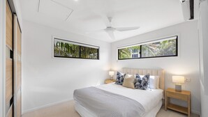 Une chambre dotée d’un grand lit, de deux tables de chevet, d’un ventilateur de plafond et de deux fenêtres donnant sur un paysage verdoyant.