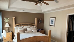 Een slaapkamer met een houten bed, een nachtkastje met een lamp, een ventilator en een klein pouf.