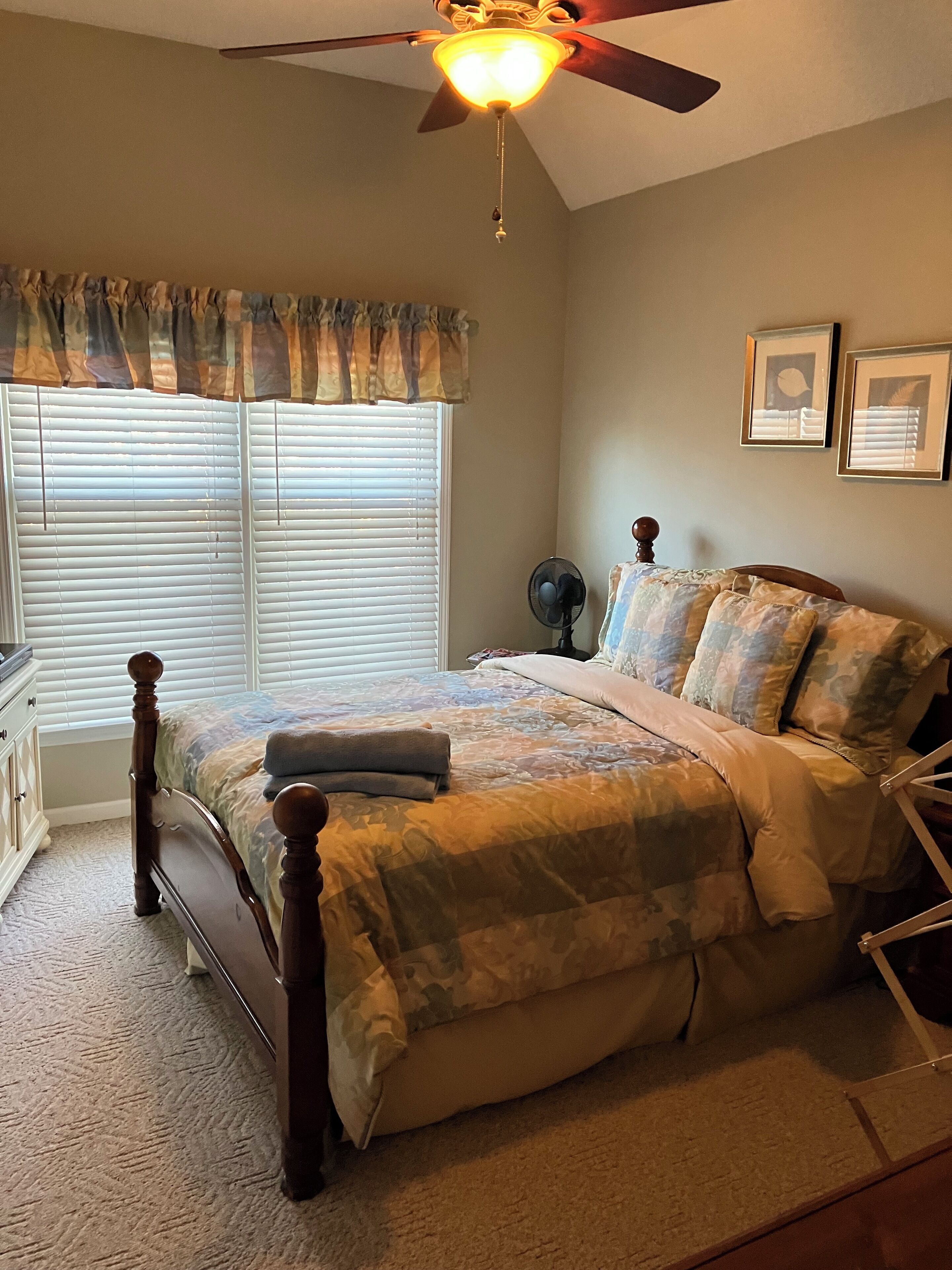 A bedroom with a bed, a ceiling fan, a window with blinds, a bedside table, and framed pictures on the wall.