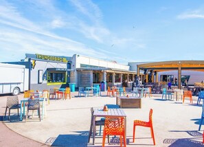 Uma área de estar ao ar livre com cadeiras e mesas coloridas, um food truck identificado como 'Thunderbird Fried Chicken' e uma área coberta para sentar ao fundo.