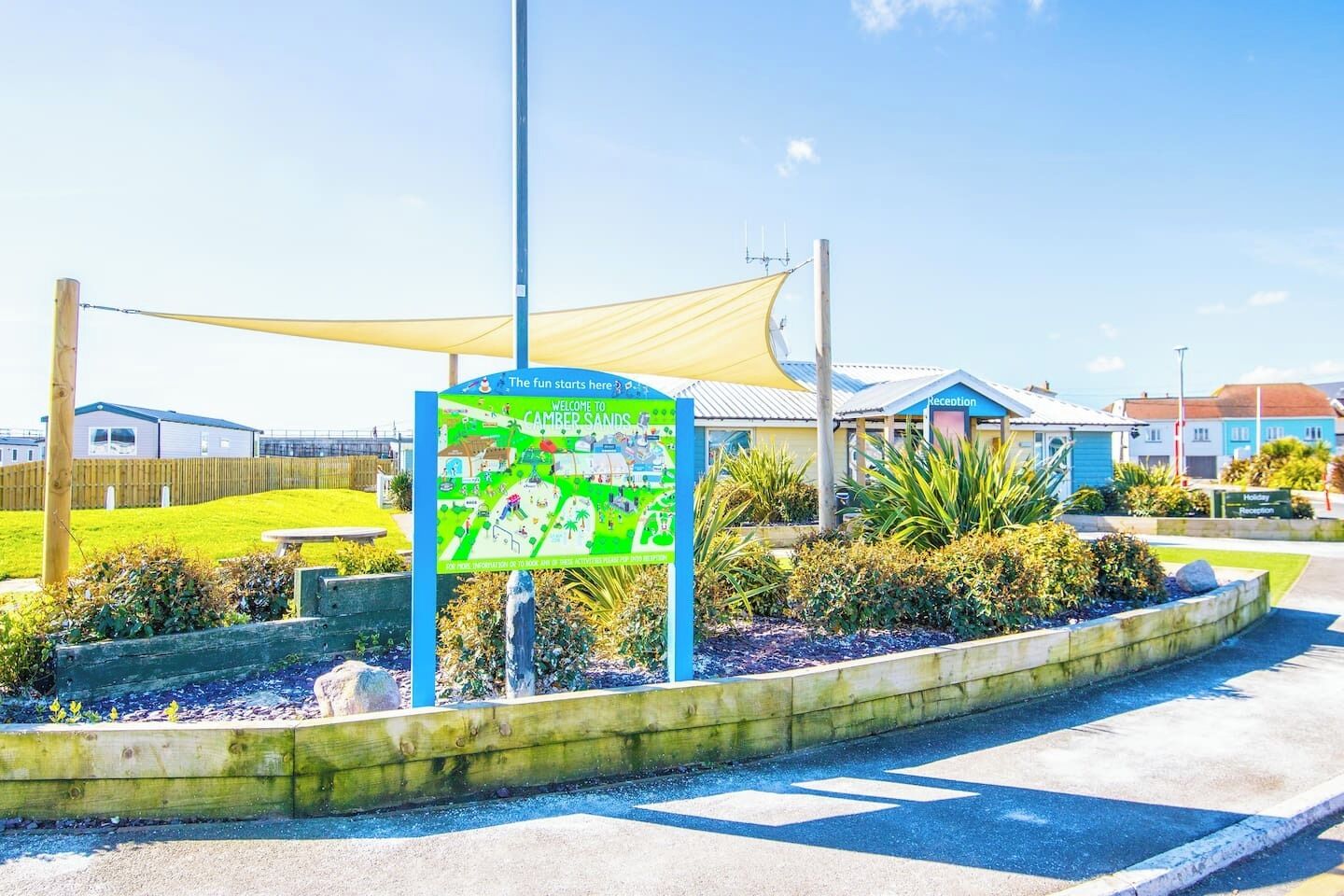 A welcome sign for Camper Sands holiday resort with a clear pathway, reception building, and surrounding greenery.