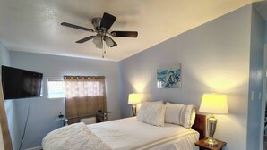Ein Hotelzimmer mit einem Bett, zwei Nachttischlampen, einem Deckenventilator, einem an der Wand montierten Fernseher und einem Fenster mit Vorhängen.