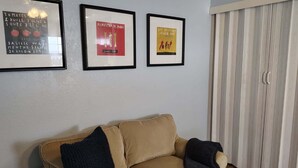 Ein Wohnzimmer mit einem beigen Sofa, drei gerahmten Bildern an der Wand und einem Fenster mit Jalousien.