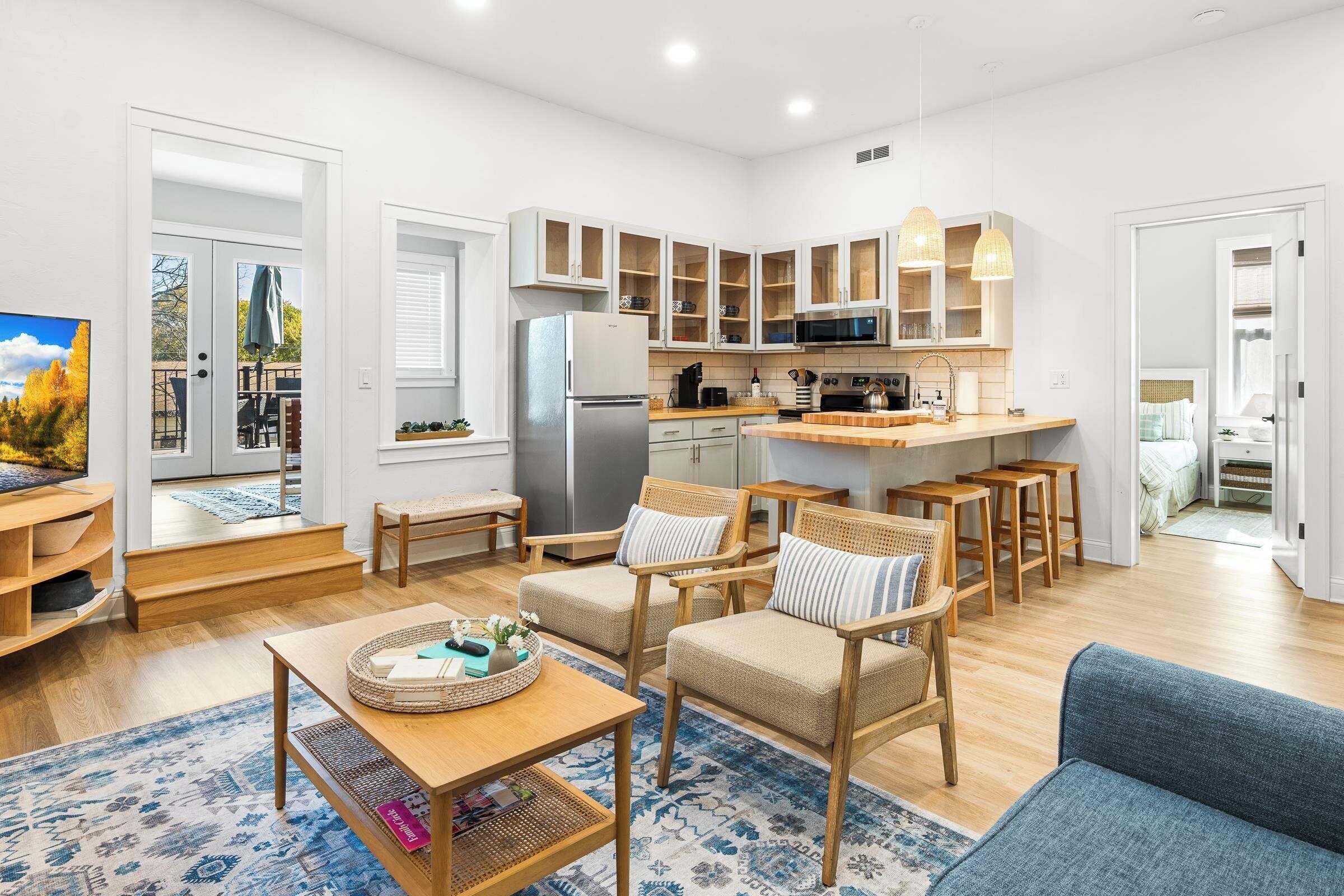 A modern kitchen with white cabinets, a refrigerator, and a microwave. There are wooden chairs and a blue sofa in the living area.