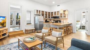 A modern kitchen with white cabinets, a refrigerator, and a microwave. There are wooden chairs and a blue sofa in the living area.