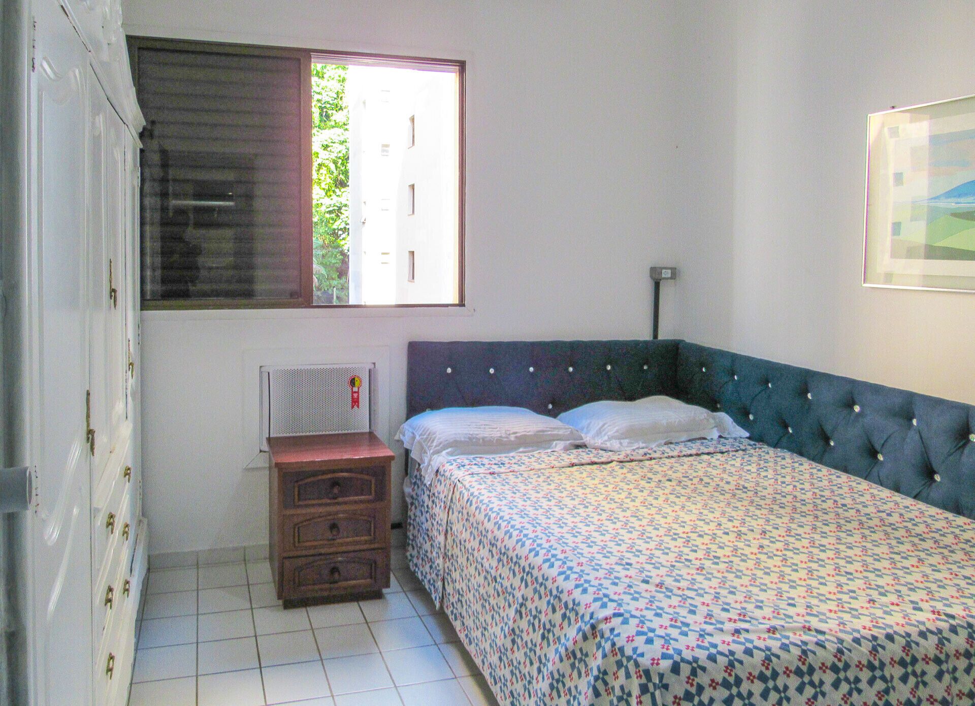 Una camera da letto con un letto, una cassettiera, un termosifone e una finestra con vista sul verde.