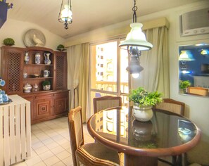 A dining area with a round table, chairs, a chandelier, a cabinet with decorative items, and a view of buildings through a window. - Apartamento a 70m da Praia de Pitangueiras/SP (Guarujá)