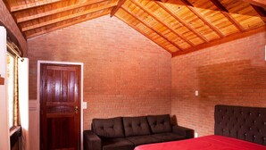 A room with a wooden ceiling, brick walls, a dark brown sofa, a red bedspread, and a wooden door.
