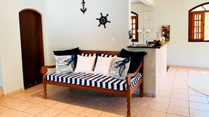 A living room with a wooden sofa, a tiled floor, a wooden door, and a kitchen area in the background. - Incrível casa a 13 min a pé da Praia de Juquehy/SP (São Sebastião)