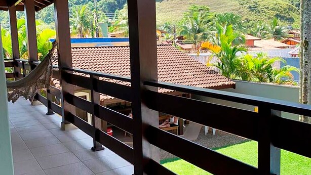 A covered balcony with a hammock, a tiled floor, and a view of a green landscape with hills and buildings. - Incrível casa a 13 min a pé da Praia de Juquehy/SP (São Sebastião)