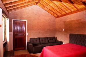 A room with a wooden ceiling, brick walls, a dark brown sofa, a red bedspread, and a wooden door.