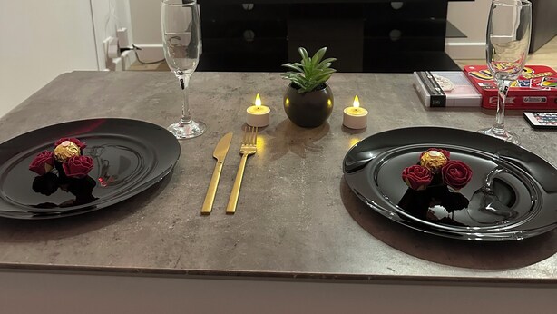 Une table à manger est dressée avec de l’argenterie dorée, des assiettes noires et des bougies. Un téléviseur, visible à l’arrière-plan, affiche le logo de Netflix.