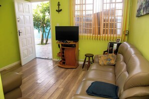 A living room with a brown leather sofa, a television on a wooden stand, and a small table with a laptop and a bag. - Casa Pet Friendly a 950m da Praia Vista Linda (Bertioga)