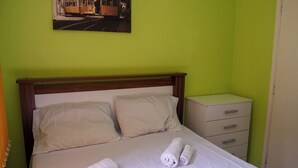 A neatly made bed with white linens and rolled towels, a wooden headboard, a nightstand with drawers, and a painting of a tram on the wall. - Casa Pet Friendly a 950m da Praia Vista Linda (Bertioga)