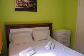 A neatly made bed with white linens and rolled towels, a wooden headboard, a nightstand with drawers, and a painting of a tram on the wall. - Casa Pet Friendly a 950m da Praia Vista Linda (Bertioga)