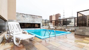 Un área de piscina en la azotea con un muro de piedra, dos sillones blancos y una vista despejada del horizonte de la ciudad.