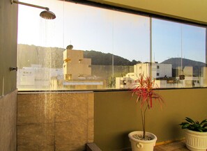A balcony with a showerhead, a potted plant, and a view of buildings and hills. - Apto a 200m Praia Pitangueiras | Piscina e Sauna (Guarujá)