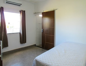 A bedroom with a bed, a window with curtains, a door, and an air conditioning unit. - Casa com Piscina e Área Gourmet | Taipu de Fora (Maraú)