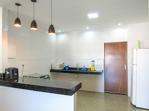 A kitchen with a black countertop, white tiles, a microwave, a sink, and a refrigerator. - Casa com Piscina e Área Gourmet | Taipu de Fora (Maraú)
