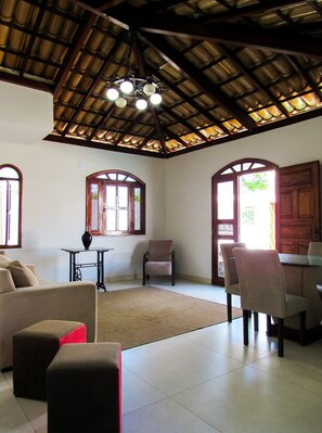 A spacious living area with a high ceiling, a dining table with chairs, a sofa, and a small table with a vase. - Casa a 250m da Praia do Sul com Wi-Fi em Ilhéus/BA (Ilhéus)