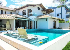 A swimming pool with lounge chairs and a sunshade, surrounded by a house with a tiled roof and a covered patio. - Casa a 250m da Praia do Sul com Wi-Fi em Ilhéus/BA (Ilhéus)