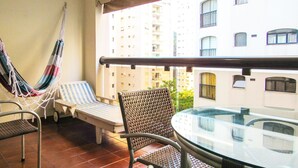 A balcony with a hammock, two lounge chairs, a table, and a view of a high-rise building. - Apto c/ Piscina e Sauna | 200m Praia Pitangueiras (Guarujá)