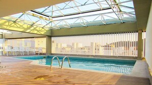 An indoor swimming pool with a wooden deck, glass ceiling, and white barriers. - Apto c/ Piscina e Sauna | 200m Praia Pitangueiras (Guarujá)
