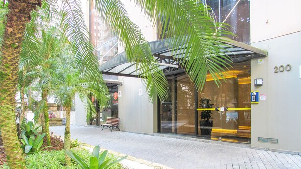 A building entrance with a glass door, a bench outside, and a number '200' on the wall. - Apto c/ Piscina e Sauna | 200m Praia Pitangueiras (Guarujá)