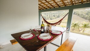 Una mesa de comedor con platos, vasos y una botella, con una hamaca y vista a un paisaje a través de una ventana.