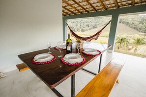 A dining table set with plates, glasses, and a bottle, with a hammock and a view of a landscape through a window. - Linda casa de campo com Wi-Fi em Santa Teresa/ES (Santa Teresa)