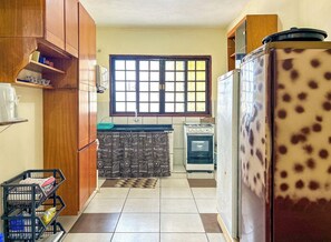 A compact kitchen with wooden cabinets, a window with blinds, a refrigerator with a spotted pattern, and a stove. - Casa c churrasqueira próx à Praia Jardim Real/SP (Praia Grande)