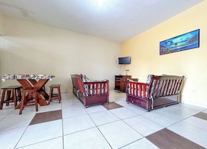 A living room with a tiled floor, a wooden dining table with chairs, two wooden sofas, a television on the wall, and a painting. - Casa c churrasqueira próx à Praia Jardim Real/SP (Praia Grande)