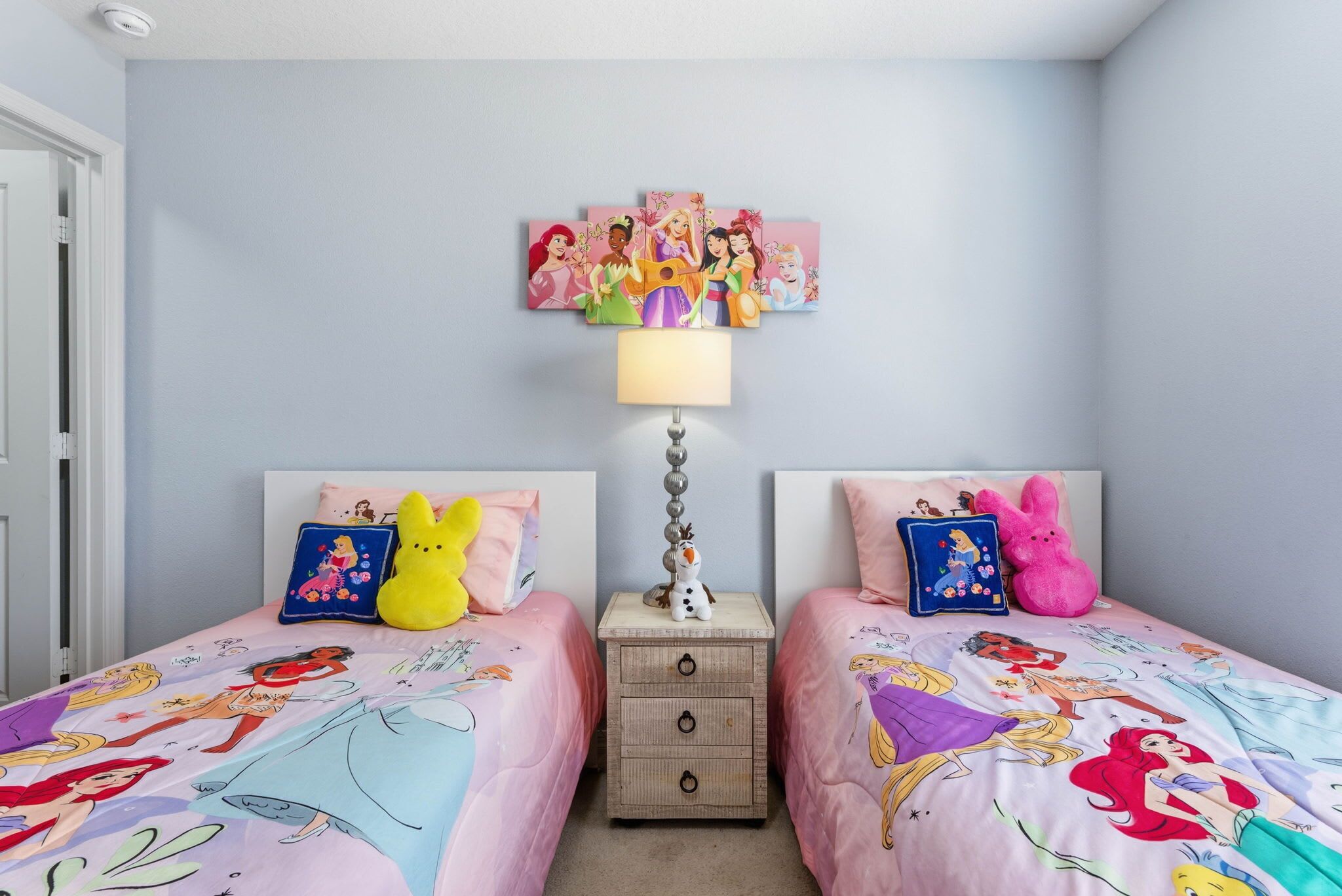 A bedroom with two beds, each with a Disney-themed bedspread, a nightstand with a lamp, and a wall-mounted Disney artwork above the beds.