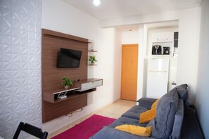 Ein Wohnzimmer mit einem an einer Holzwand montierten Flachbildfernseher, einem grauen Sofa mit gelben Kissen, einem rosa Teppich und einer Holztür im Hintergrund.