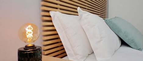 A modern bedroom with a wooden slatted headboard, a bedside table with a glass bulb lamp, and white bedding.