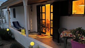 Un espacio al aire libre cubierto con mesa y sillas, una planta en maceta y la entrada de un edificio con puerta y ventana.