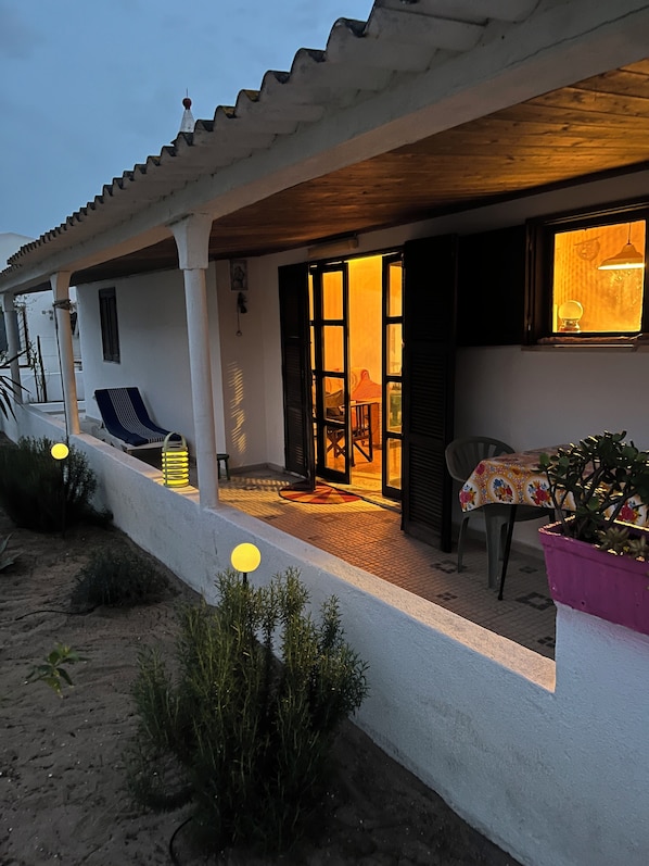Un espace extérieur abrité, aménagé avec une table et des chaises, un pot de fleurs et l’entrée d’un bâtiment, avec une porte et une fenêtre.