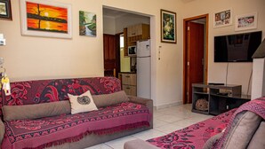 A living room with a sofa, a TV, and a kitchen in the background. - Casa c ótima localização e churrasq em Mongaguá/SP (Mongaguá)