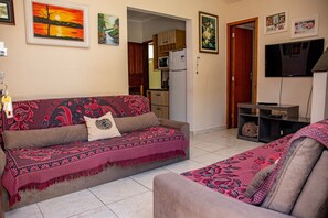 A living room with a sofa, a TV, and a kitchen in the background. - Casa c ótima localização e churrasq em Mongaguá/SP (Mongaguá)