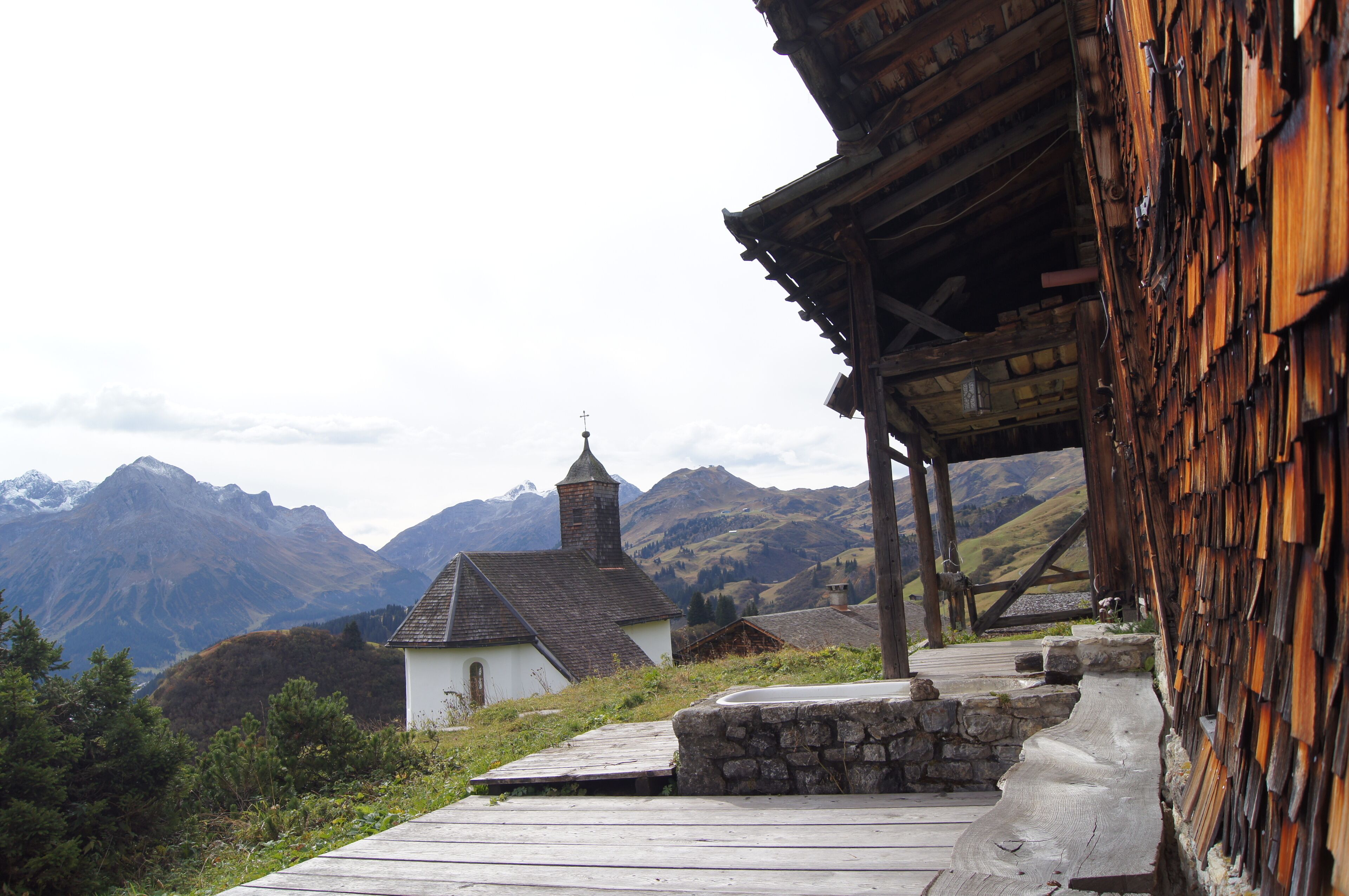 Un bâtiment rustique en bois, avec un chemin de pierre menant à une petite église blanche dotée d’un clocher, le tout entouré de montagnes.