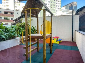 Eine Spielanlage aus Holz mit Rutsche und kleinem Tisch, umgeben von Blumentöpfen und bunten Fliesen auf einer Dachterrassen.
