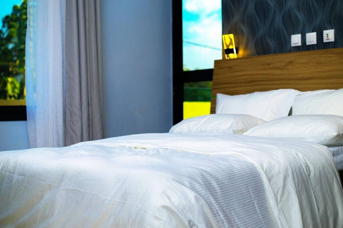 A bed with white bedding in a room with a wooden headboard, a window with curtains, and a wall with a textured pattern.