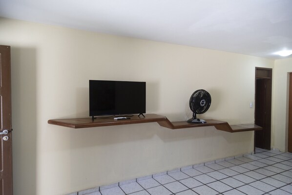 A room with a television mounted on a wooden shelf and a standing fan on the floor.