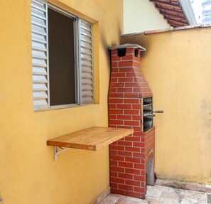 A brick-built outdoor oven attached to a yellow wall, with a window and a wooden shelf below. - Casa aconchegante c churrasqueira à 400m da praia (Praia Grande)