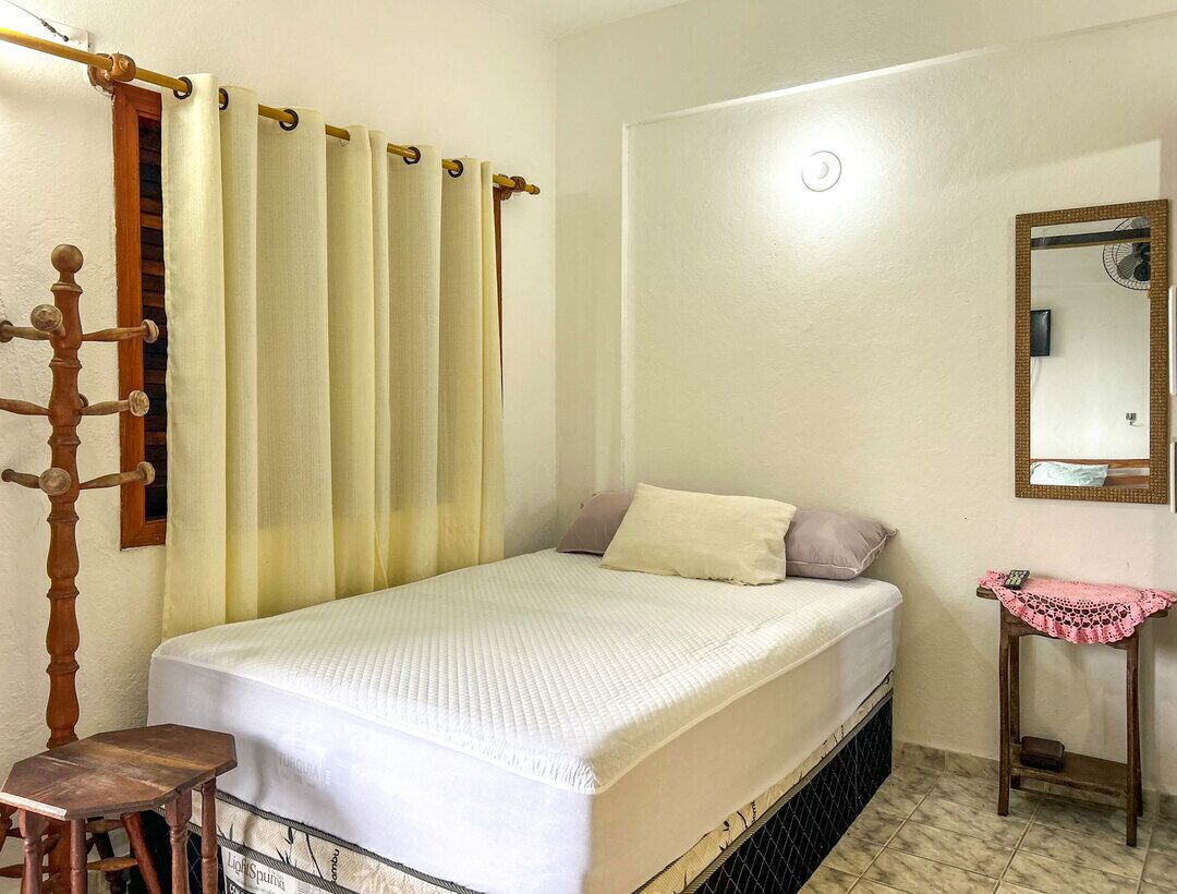 Una habitación pequeña con cama individual, estructura de cama de madera, mesita de noche con un paño rosa, un espejo en la pared y una barra con cortina.